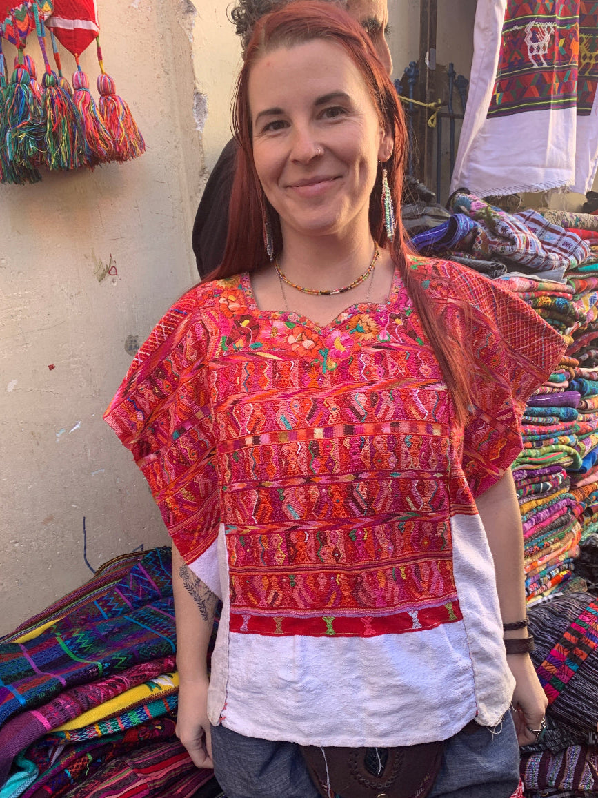 Woman wearing a colorful red and white hüipil in a market setting with various textiles.