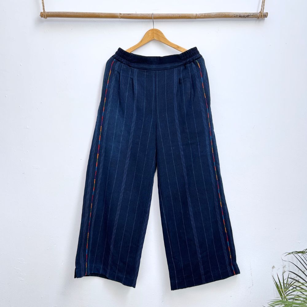 Navy pinstripe corte trousers with thin colorful randa on the sides, hanging on a wooden hanger against a white background.