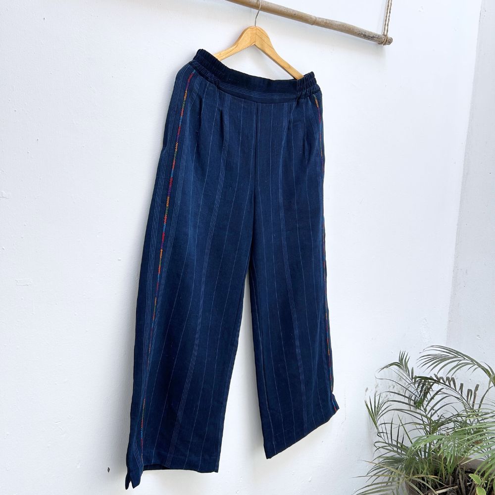 Navy pinstripe corte trousers with thin colorful randa on the sides, hanging on a wooden hanger against a white background.