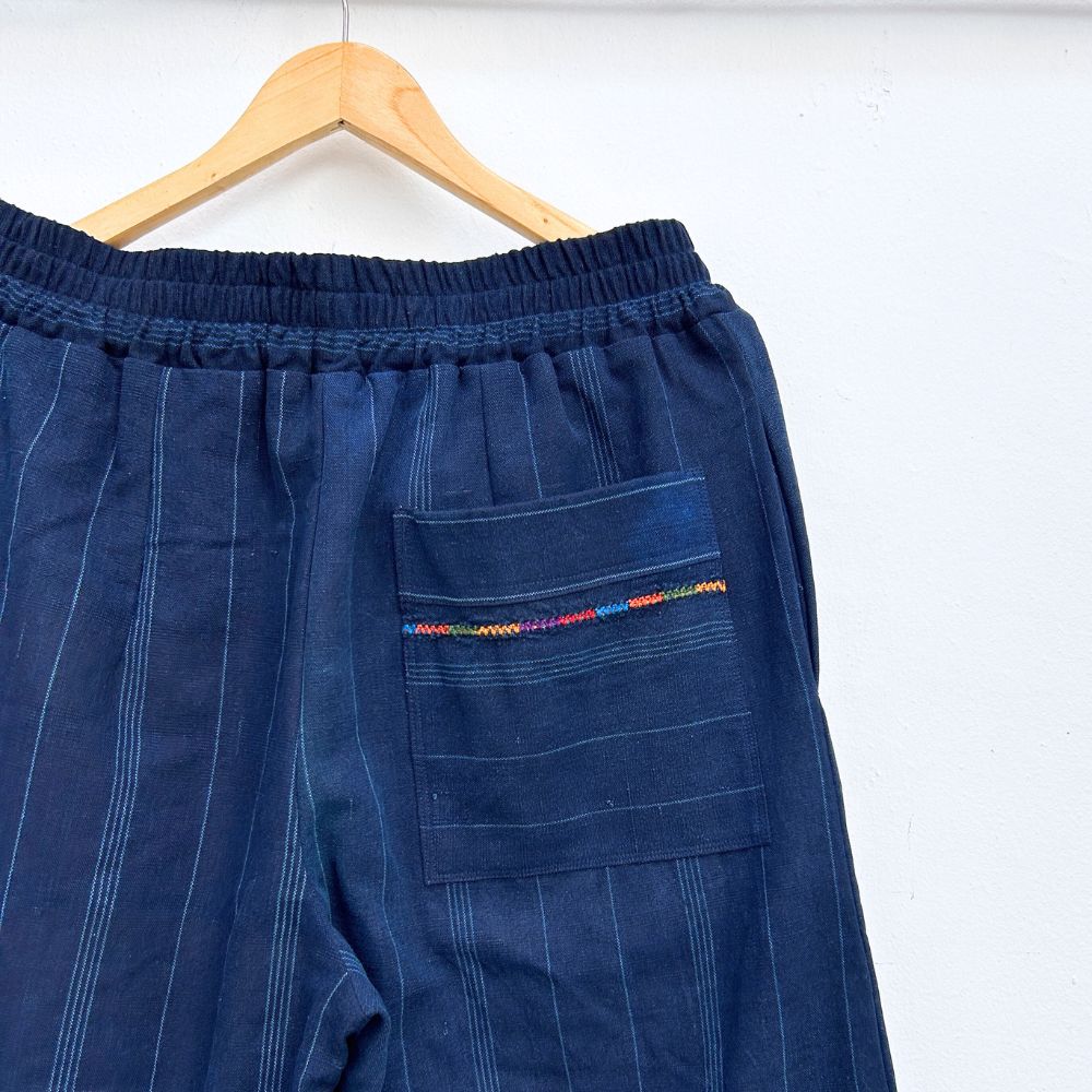 Navy pinstripe corte trousers with thin colorful randa on the sides, hanging on a wooden hanger against a white background.
