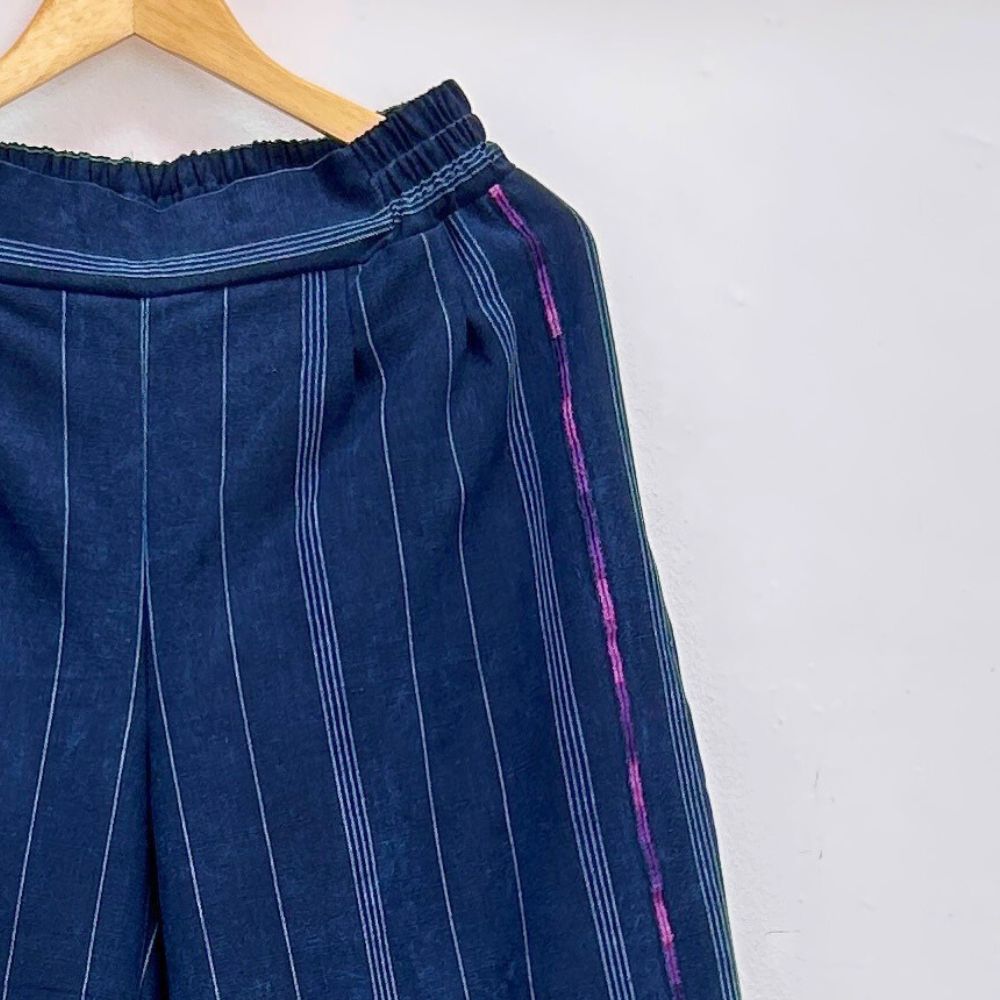 Blue pinstripe pants with pink and purple randa embroidery stitching on a hanger against a white background