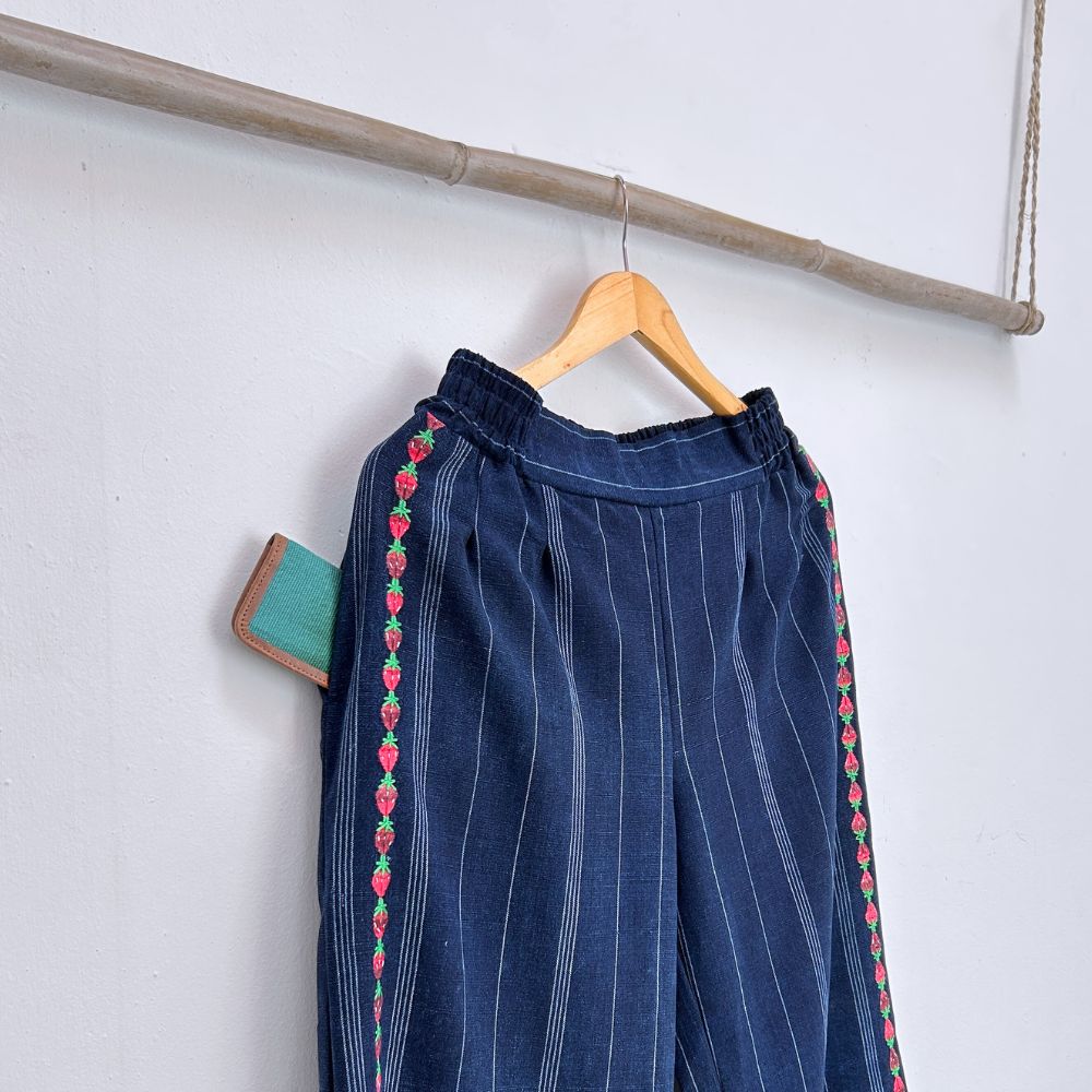 Blue pants with red and green strawberries embroidery on a wooden hanger against a white wall.