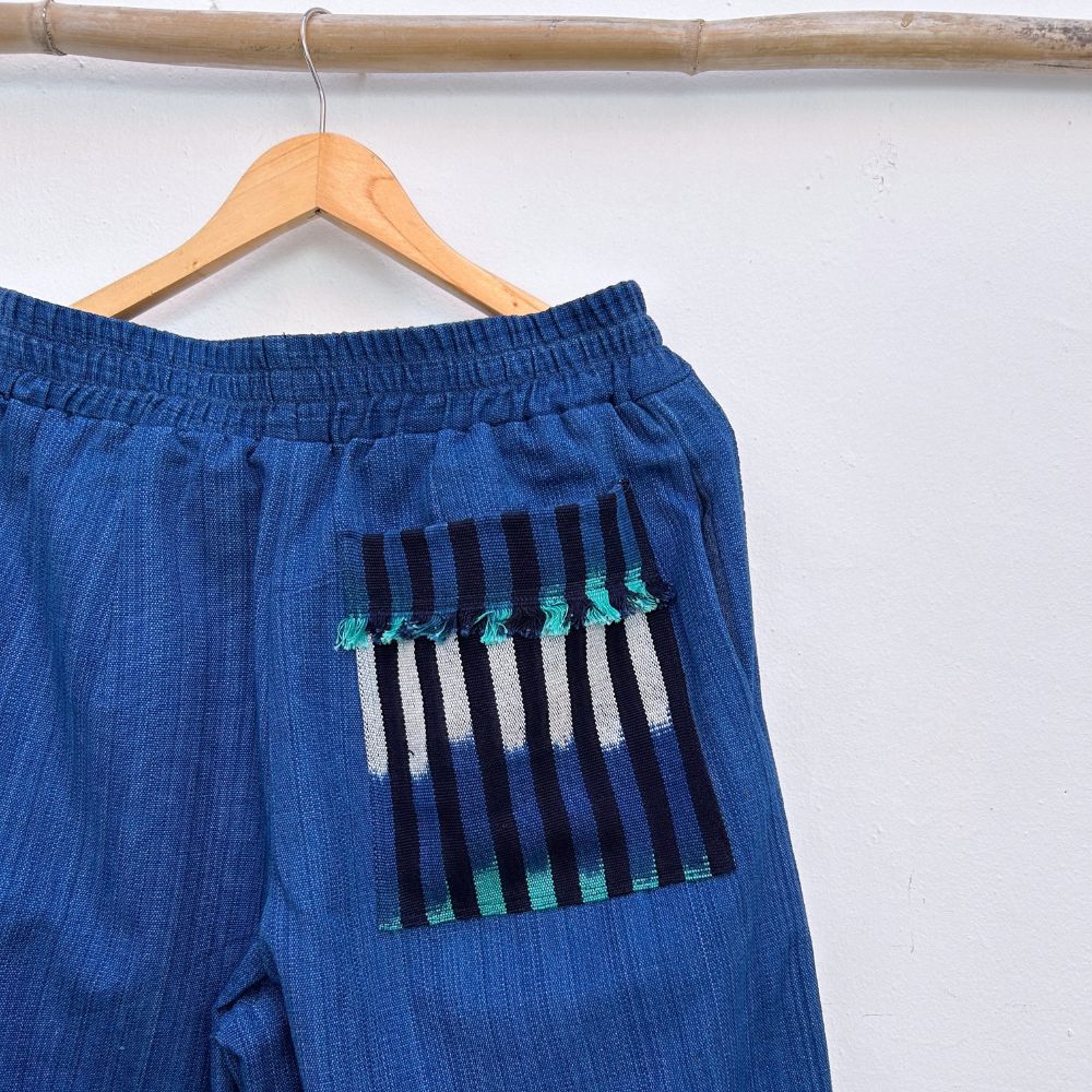 Blue pants with a striped pocket on a wooden hanger against a white wall.