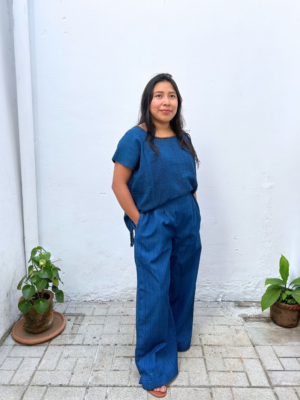 Woman in a blue outfit standing against a white wall with plants on either side.