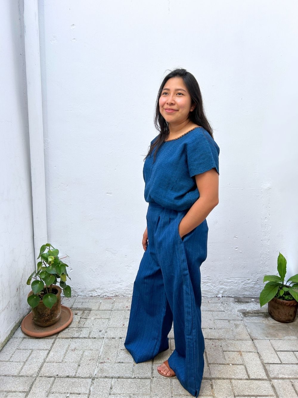 Woman in a blue outfit standing against a white wall with plants around.
