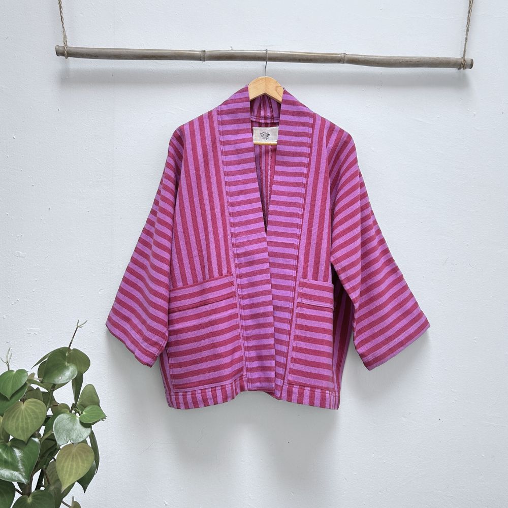Pink and purple striped jacket hanging on a wooden hanger against a white wall with a plant on the left.