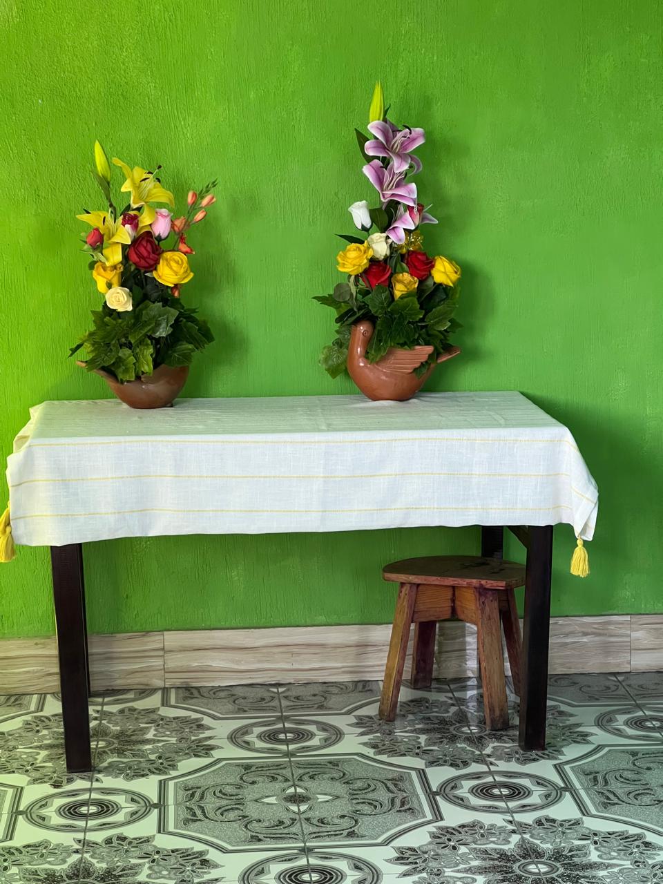 A white tablecloth with yellow stripes and tassel ties is draped over a dark wooden table, holding two terracotta flower pots with colorful flowers. The setup is against a bright green wall and a patterned gray tile floor, with a small wooden stool underneath.