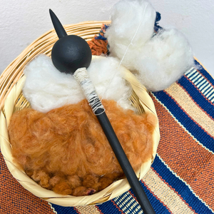 A handcrafted cotton spinning setup featuring a black wooden spindle with a wrapped handle, resting in a woven straw basket filled with brown cotton (ixcaco) fibers and white wool roving, set on a striped textile.