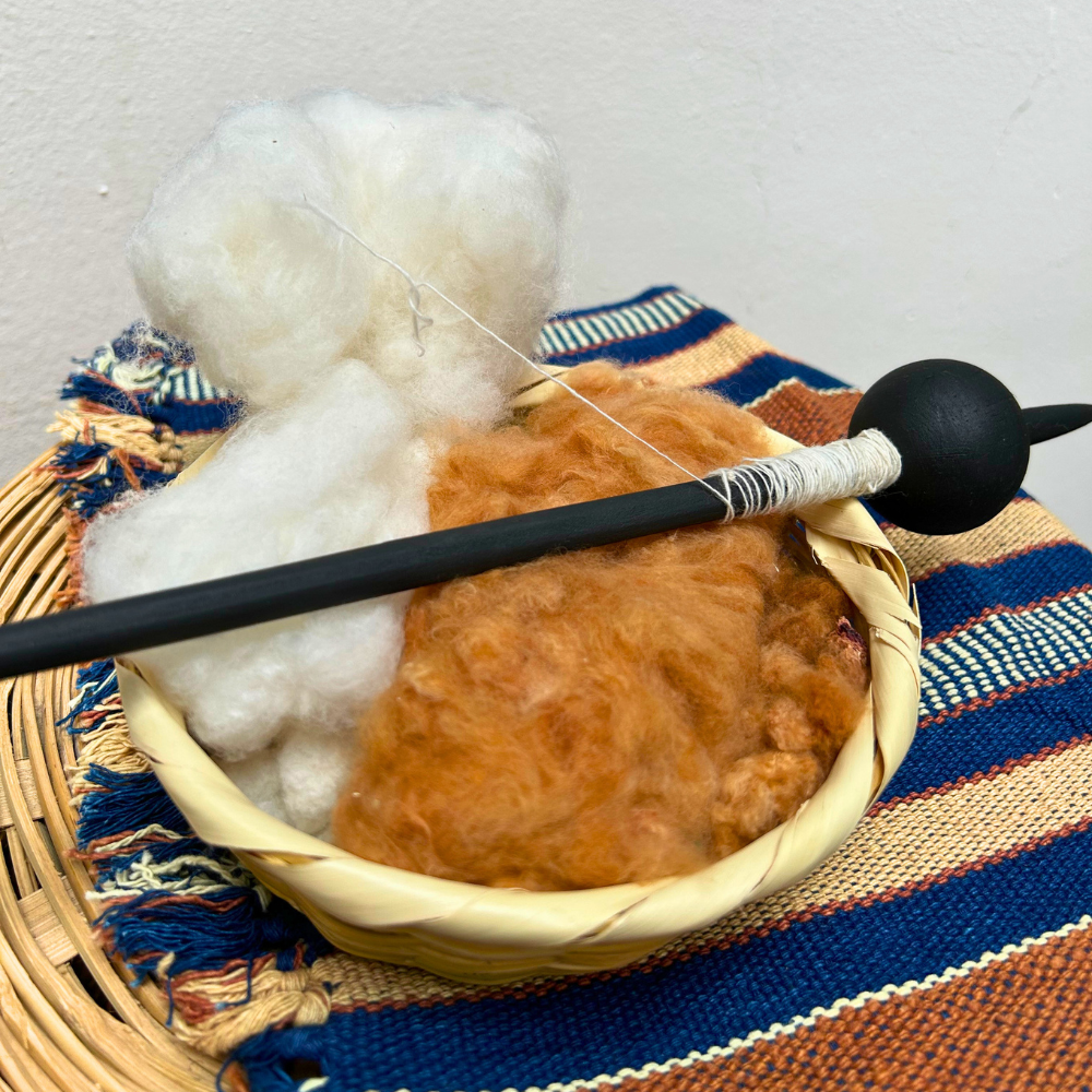 A close up of a spinning wheel setup with a black spindle resting across a woven basket filled with white cotton fiber and brown cotton, placed on a striped textile.