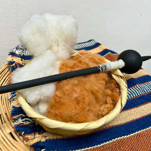 A close up of a spinning wheel setup with a black spindle resting across a woven basket filled with white cotton fiber and brown cotton, placed on a striped textile.