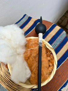 A close up of a handcrafted fiber art piece in a woven basket, featuring fluffy white and brown cotton fibers with a black pointed tool resting on top, set against a striped woven textile.