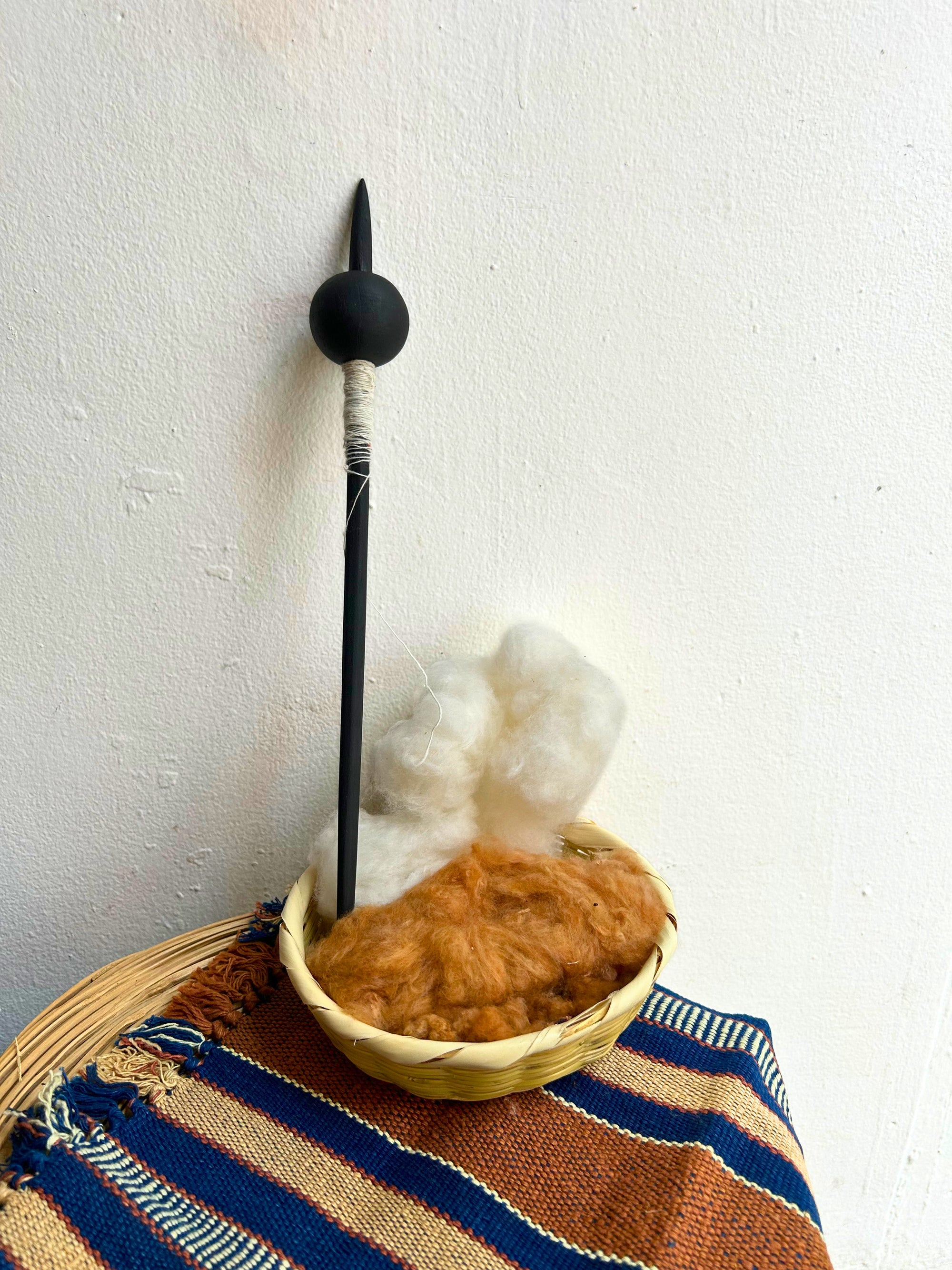 A black Malacate and white wrapped shaft stands upright in a small woven basket filled with fluffy white and brown cotton, set on a striped woven textile against a plain white wall.