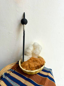 A black Malacate and white wrapped shaft stands upright in a small woven basket filled with fluffy white and brown cotton, set on a striped woven textile against a plain white wall.
