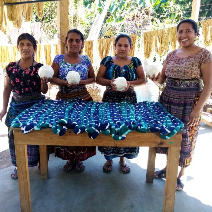 Four women in traditional clothing holding white balls of yarn behind a table with ikat-dyed threads.