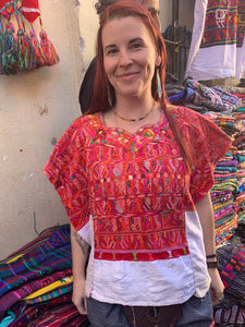 Woman wearing a colorful red and white hüipil in a market setting with various textiles.