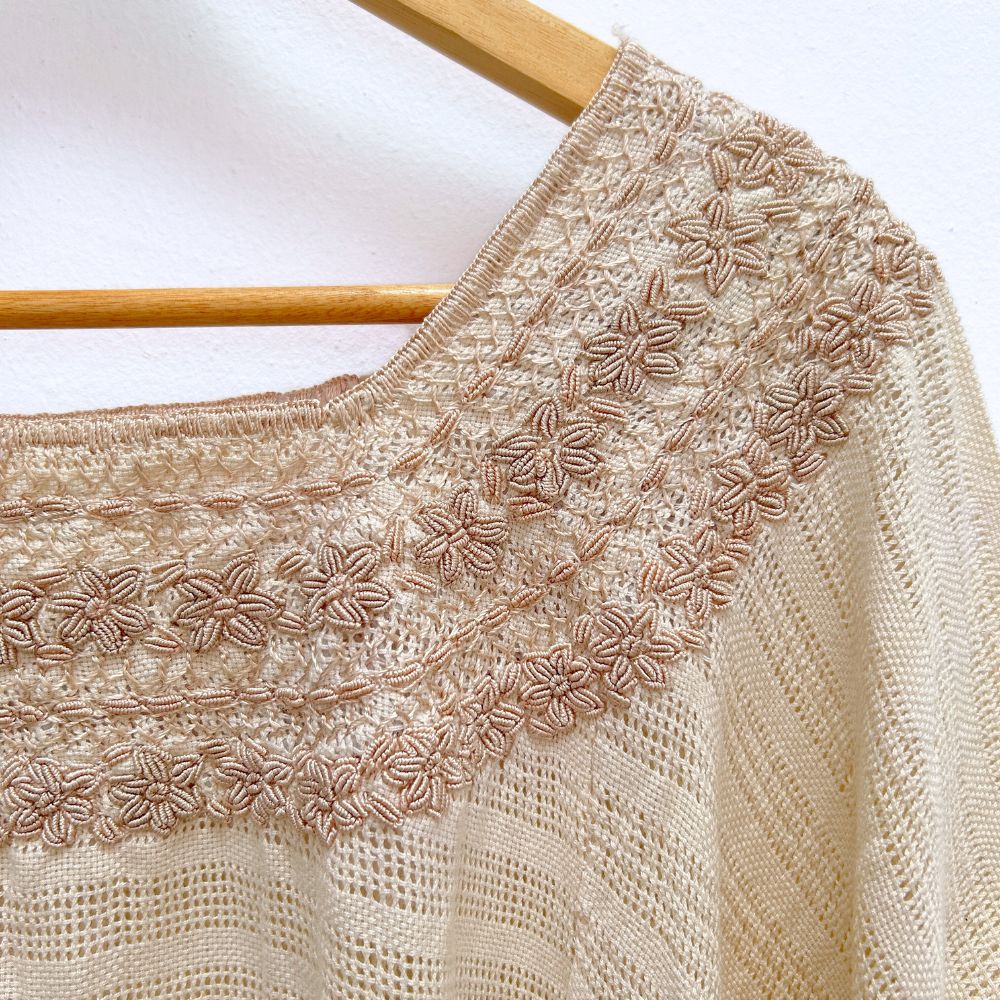 Beige base woven blouse with embroidery french knot neckline  same color on a wooden hanger against a white wall.