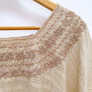Beige base woven blouse with embroidery french knot neckline  same color on a wooden hanger against a white wall.