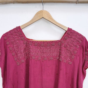 Magenta base woven blouse with embroidery french knot neckline   on a wooden hanger against a white wall.