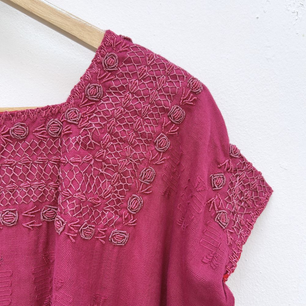 Magenta base woven blouse with embroidery french knot neckline   on a wooden hanger against a white wall.