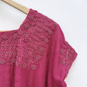 Magenta base woven blouse with embroidery french knot neckline   on a wooden hanger against a white wall.