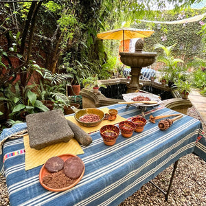 Outdoor setting with a table covered by a blue striped tablecloth, food items, and a fountain in the background.