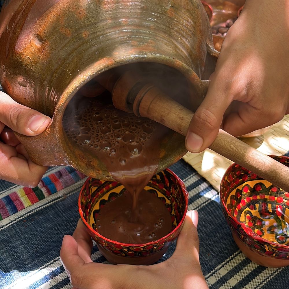 Person using a traditional wooden mortar and pestle to grind ingredients in a pot, with a colorful bowl on a striped fabric background.