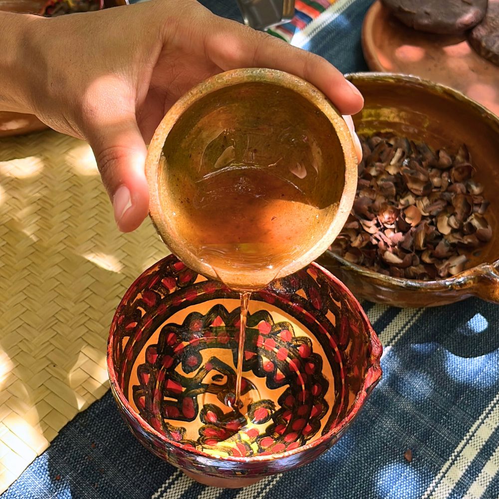 Hand pouring honey from a small bowl into a larger decorated bowl on a textured surface.