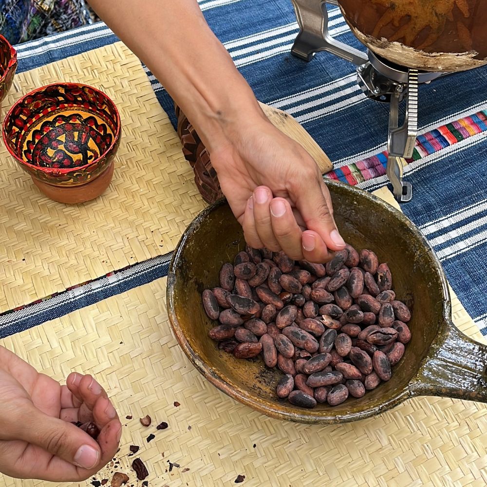 Person handling cocoa beans in a rustic setting with a woven surface and colorful background.