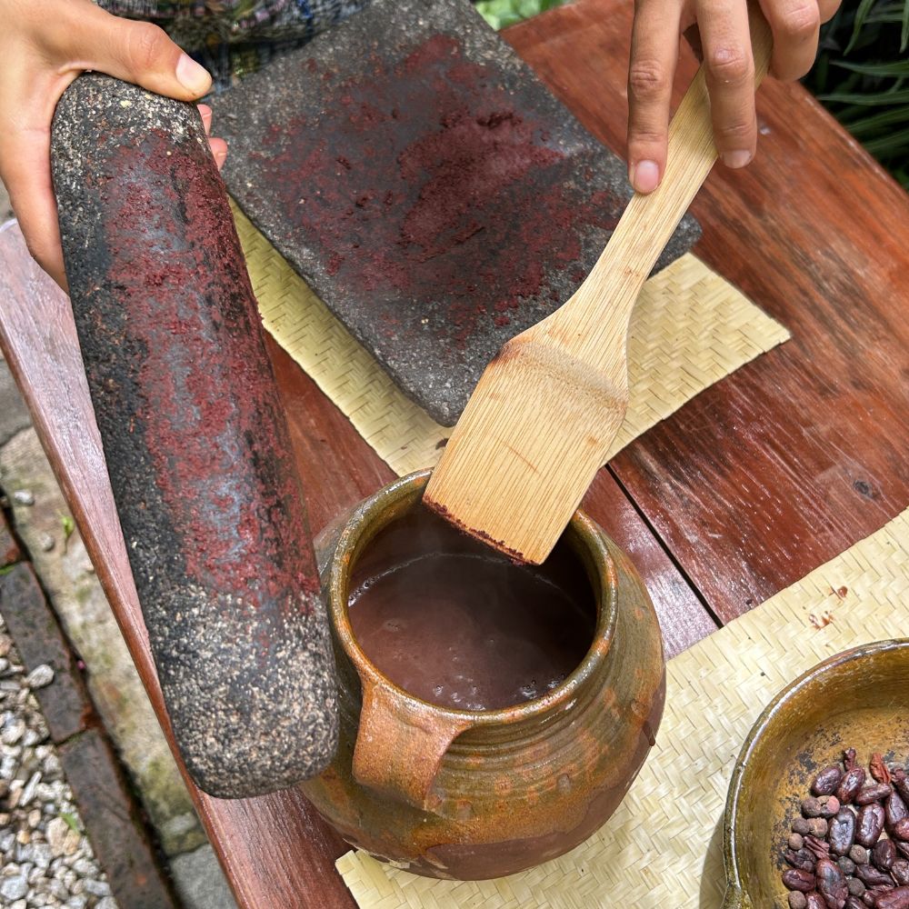 Person using a wooden spatula to scoop dark liquid from a ceramic pot on a rustic wooden surface.
