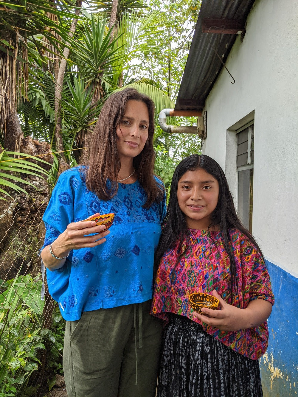Two women wearing traditional huipiles and drinking hot cacao from painted gourds.