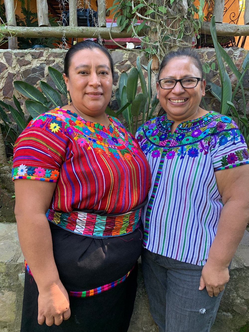 Two women standing outdoors, wearing traditional, colorful, embroidered clothing.