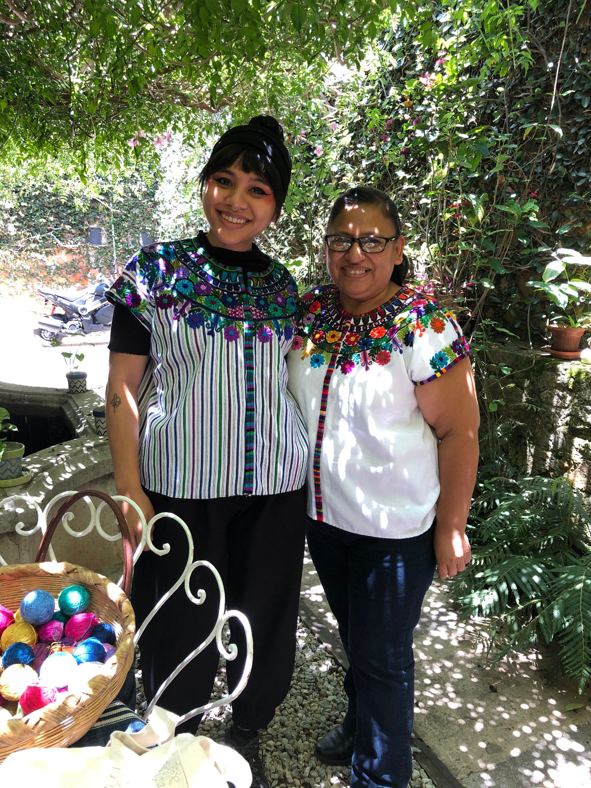 Two people in traditional embroidered huipiles standing outdoors with trees and a fountain in the background.