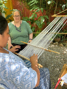 Two women working with a loom outdoors, surrounded by greenery.