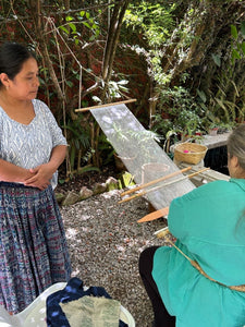 Two women in a garden setting with one weaving on a loom.