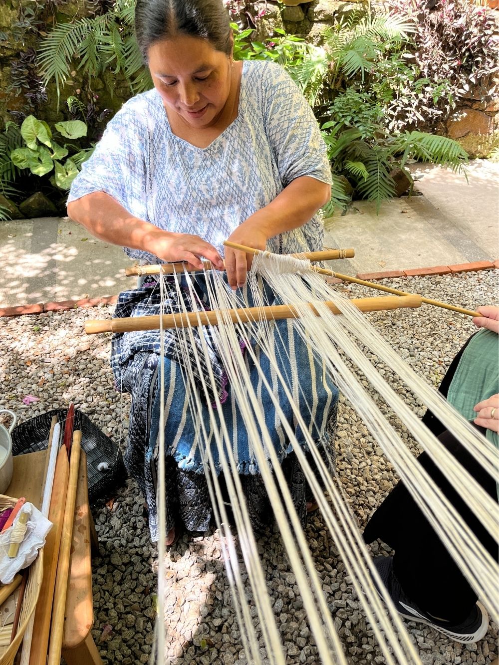 Woman weaving with yarn outdoors surrounded by plants