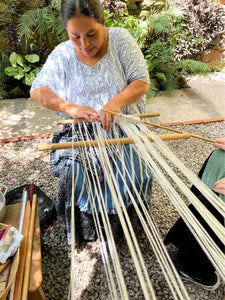 Woman weaving with yarn outdoors surrounded by plants