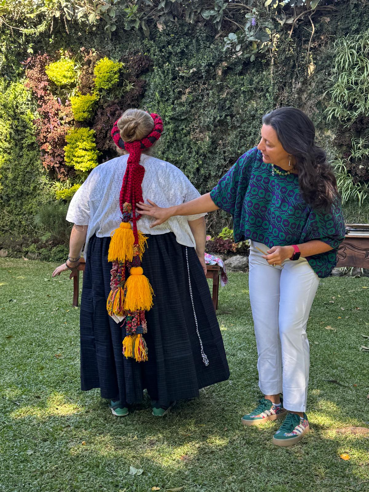 Two women, one in traditional attire with colorful hair ribbon and another in a patterned top and white pants, standing outdoors.