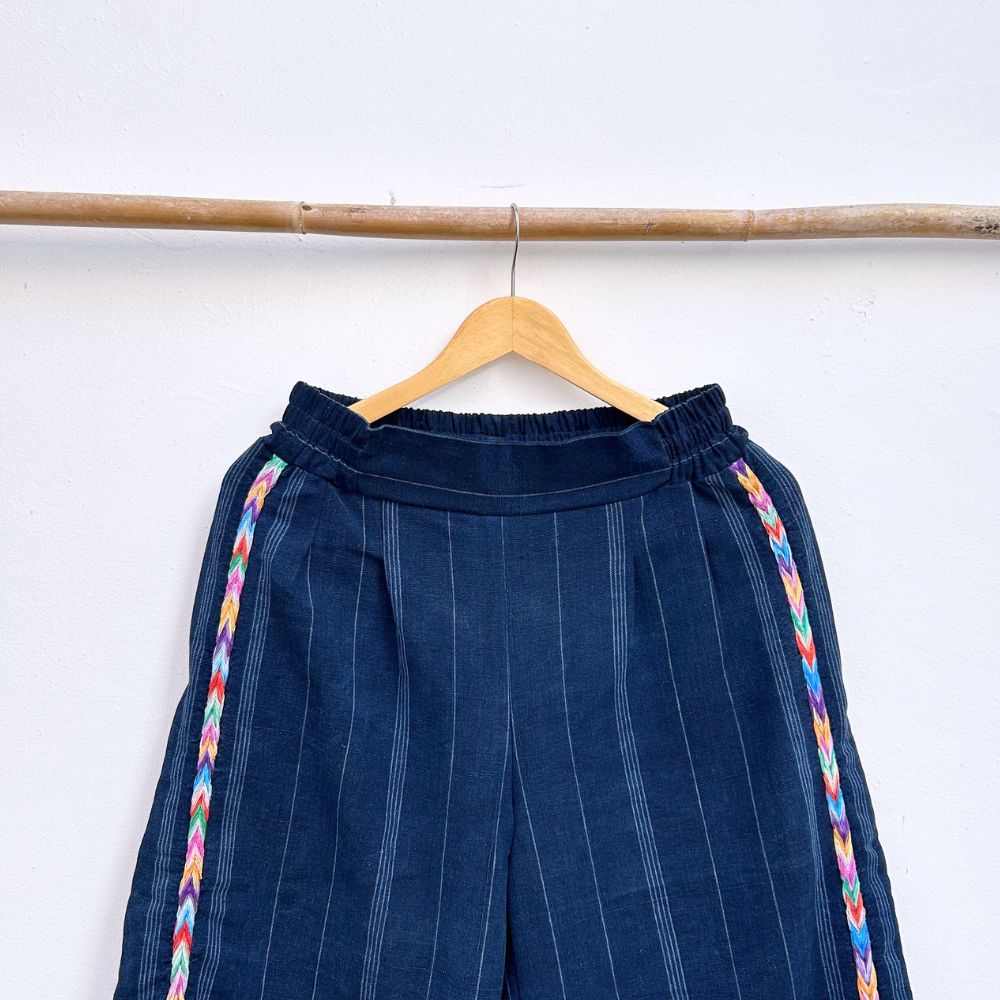 Navy pinstripe corte trousers with colorful randa on the sides, hanging on a wooden hanger against a white background.