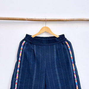 Navy pinstripe corte trousers with colorful randa on the sides, hanging on a wooden hanger against a white background.