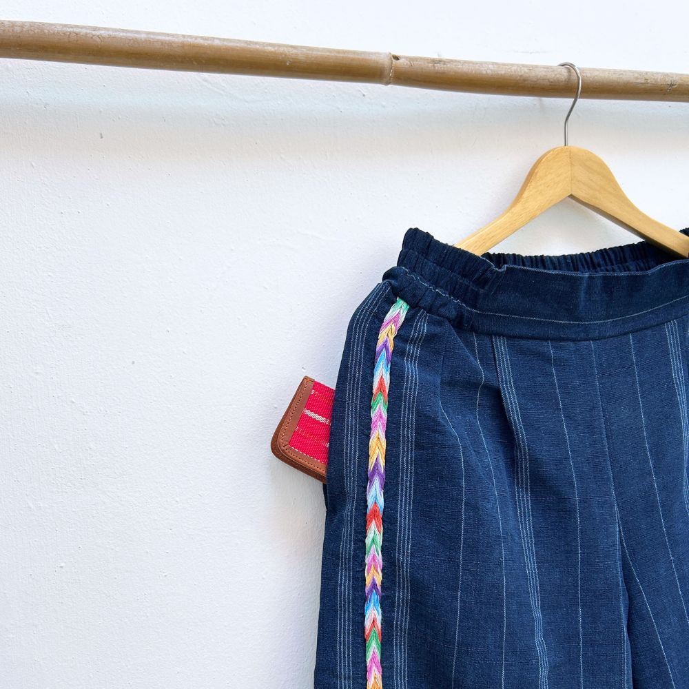 Navy pinstripe corte trousers with colorful randa on the sides, hanging on a wooden hanger against a white background.