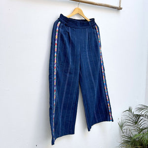 Navy pinstripe corte trousers with colorful randa on the sides, hanging on a wooden hanger against a white background.