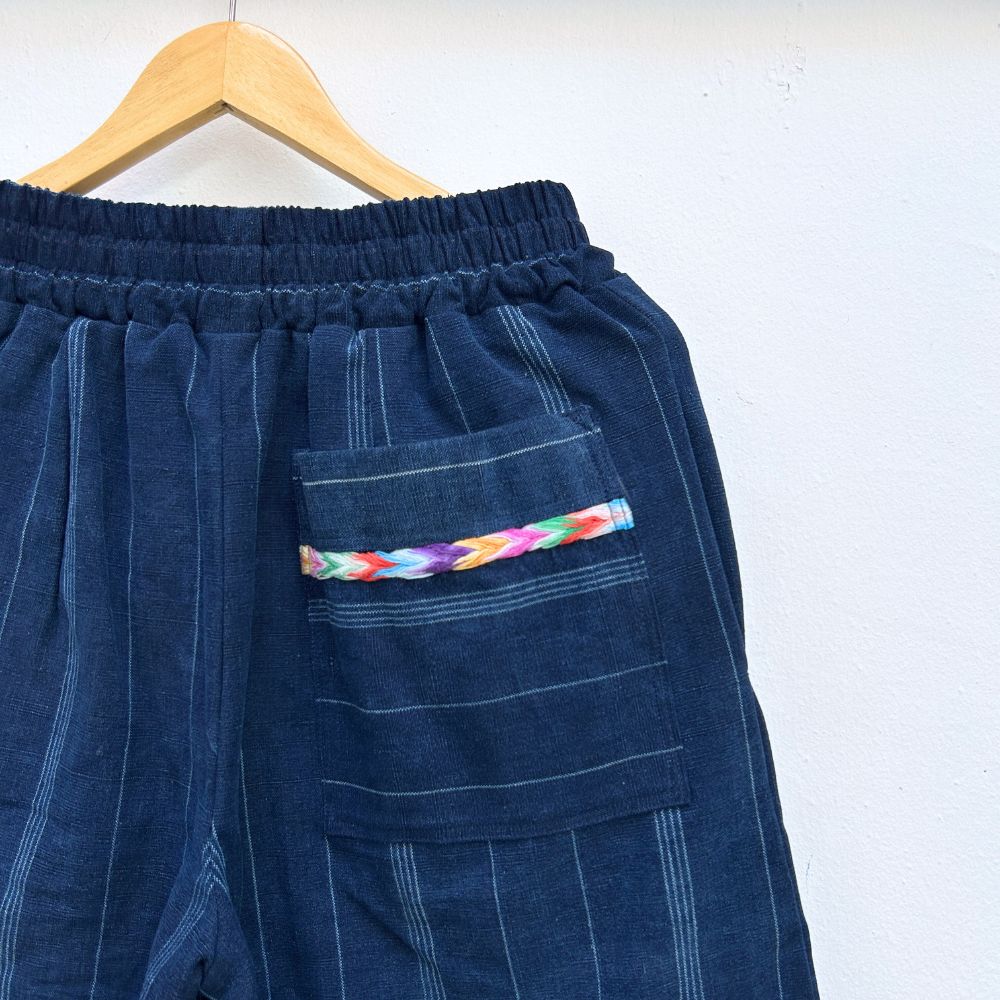 Navy pinstripe corte trousers with colorful randa on the sides, hanging on a wooden hanger against a white background.