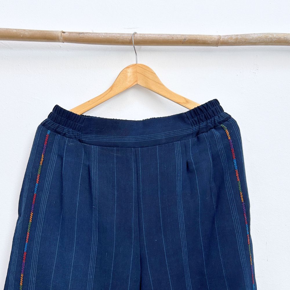 Navy pinstripe corte trousers with thin colorful randa on the sides, hanging on a wooden hanger against a white background.