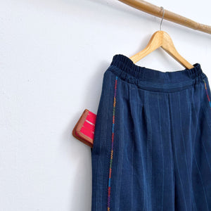 Navy pinstripe corte trousers with thin colorful randa on the sides, hanging on a wooden hanger against a white background.