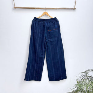 Navy pinstripe corte trousers with thin colorful randa on the sides, hanging on a wooden hanger against a white background.