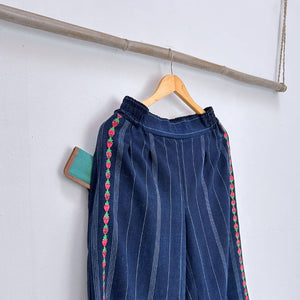 Blue pants with red and green strawberries embroidery on a wooden hanger against a white wall.