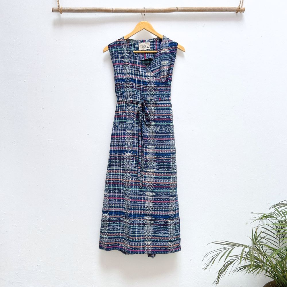 Blue patterned dress hanging on a wooden hanger against a white wall with a plant in the corner.