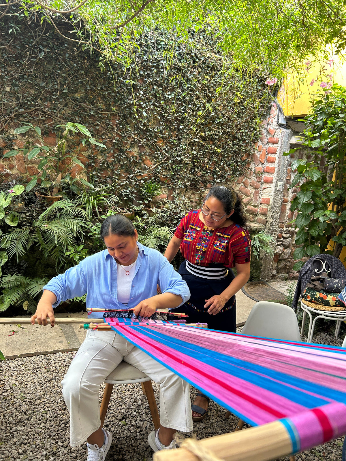 Teacher overlooking as student weaves on a colorful backstrap loom outdoors.