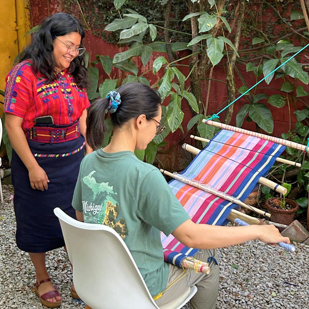 A weaving teacher guiding a student through traditional backstrap loom weaving techniques.
