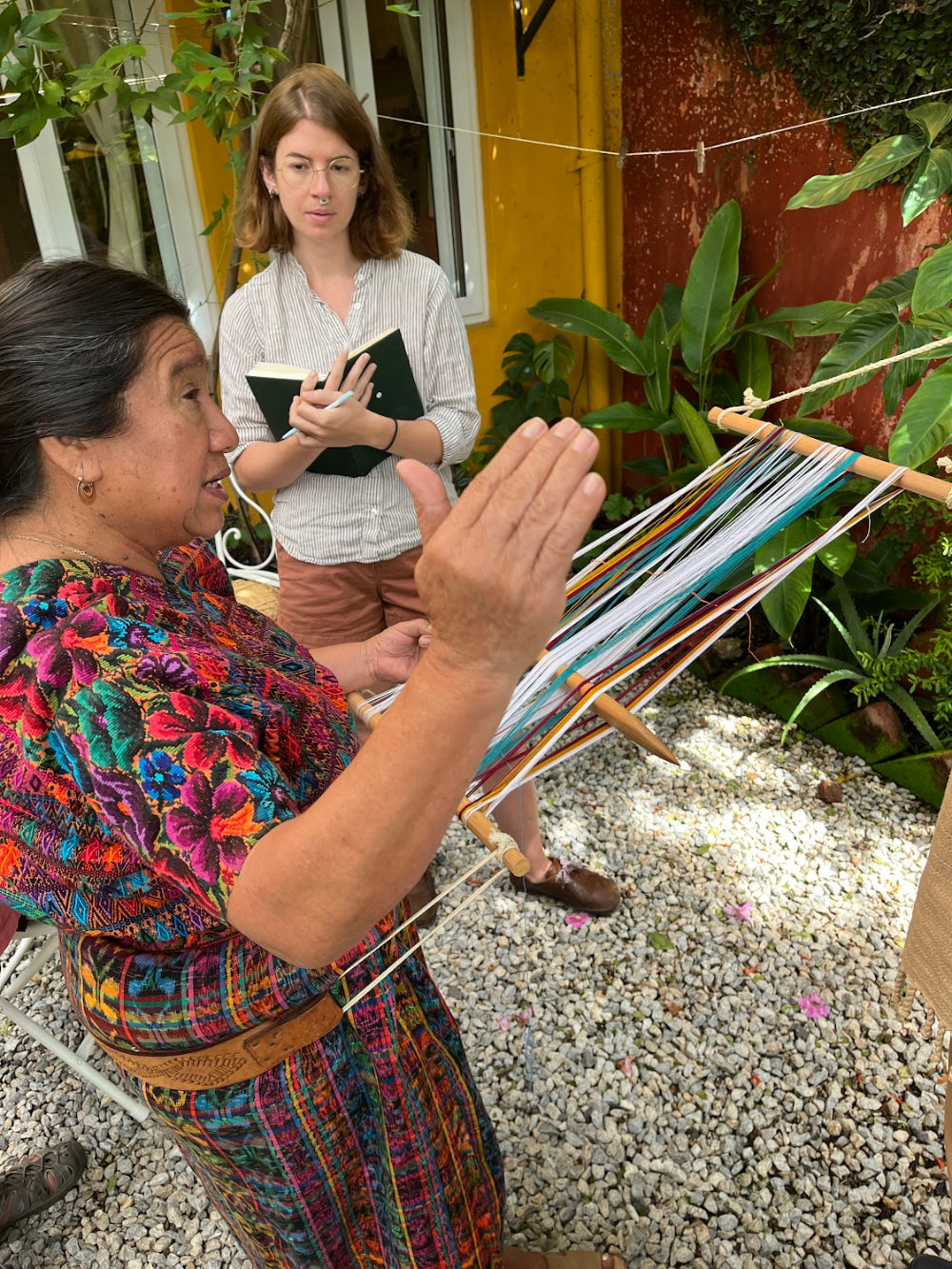 Master weaver in traditional dress weaving on a loom outdoors, with another person observing and taking notes.